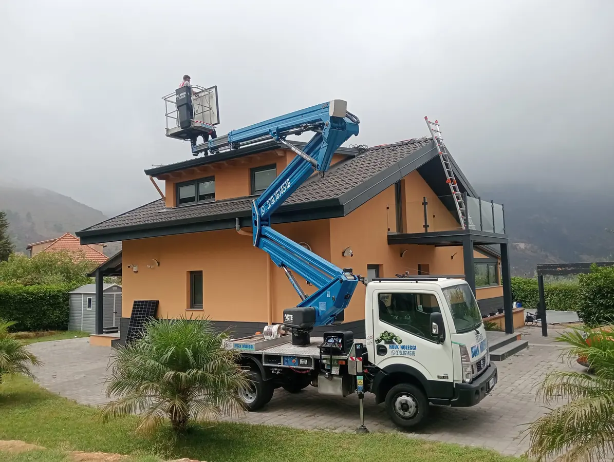 Camion col braccio blu davanti a una villetta, cestello all'altezza del tetto, montagne nella nebbia
