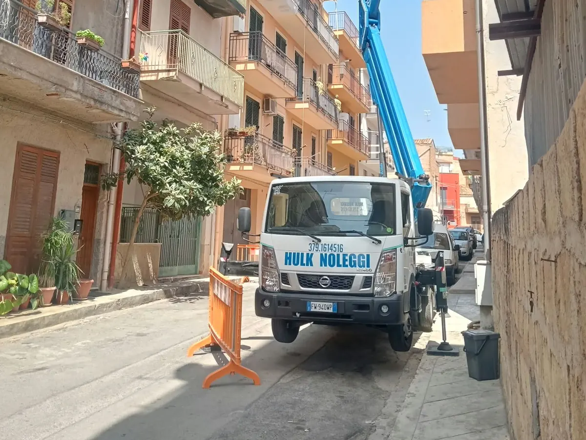 Il camion Hulk Noleggi col braccio blu in una via stretta fra le palazzine, transenna arancione
